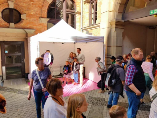 Fotobox auf dem Stadtfest am Rathaus Wiesbaden Eine Familie posiert für ein Foto in einer Fotobox, die unter einem weißen Zelt auf dem Stadtfest Wiesbaden am Rathaus aufgestellt ist. Im Vordergrund warten weitere Besucher in einer Schlange, um ebenfalls ein Foto zu machen.