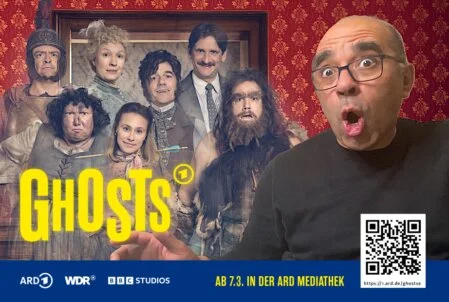 Fotobox-Layout der Serie "Ghosts" mit einem Besucher Ein Mann mit Brille macht ein überraschtes Gesicht, während er für ein Fotobox-Foto mit dem "Ghosts"-Plakat der ARD posiert. Das Plakat zeigt die Charaktere der Serie im Hintergrund.