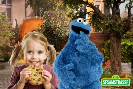 Fotobox-Spaß mit Krümelmonster und Sesamstraße-Overlay Ein lächelndes kleines Kind mit Zöpfen und einem großen Schokoladenkeks posiert neben der Figur des Krümelmonsters. Das Foto ist mit dem Logo der Sesamstraße versehen.