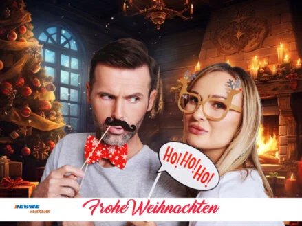 Fotobox-Spaß auf der Weihnachtsfeier Ein Mann und eine Frau posieren mit weihnachtlichen Requisiten vor einem KI-generierten Kamin-Hintergrund und haben Spaß an der Fotobox auf einem Firmenevent. Das Foto ist mit einem ESWE Verkehr Logo und einem "Frohe Weihnachten"-Schriftzug versehen.