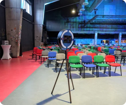 Fotobox bereit für den Einlass beim Firmenevent Eine Fotobox steht im Veranstaltungsraum eines Firmenevents, kurz bevor die Gäste eintreffen. Im Hintergrund sind Reihen von bunten Stühlen zu sehen, die auf die Veranstaltung warten.
