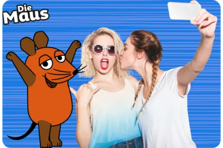 Partygäste machen ein Selfie mit der Maus-Fotobox Zwei junge Frauen posieren lachend für ein Selfie. Eine der Frauen trägt eine Sonnenbrille und macht einen lustigen Ausdruck. Die Figur "Die Maus" ist als Fotobox-Overlay sichtbar.