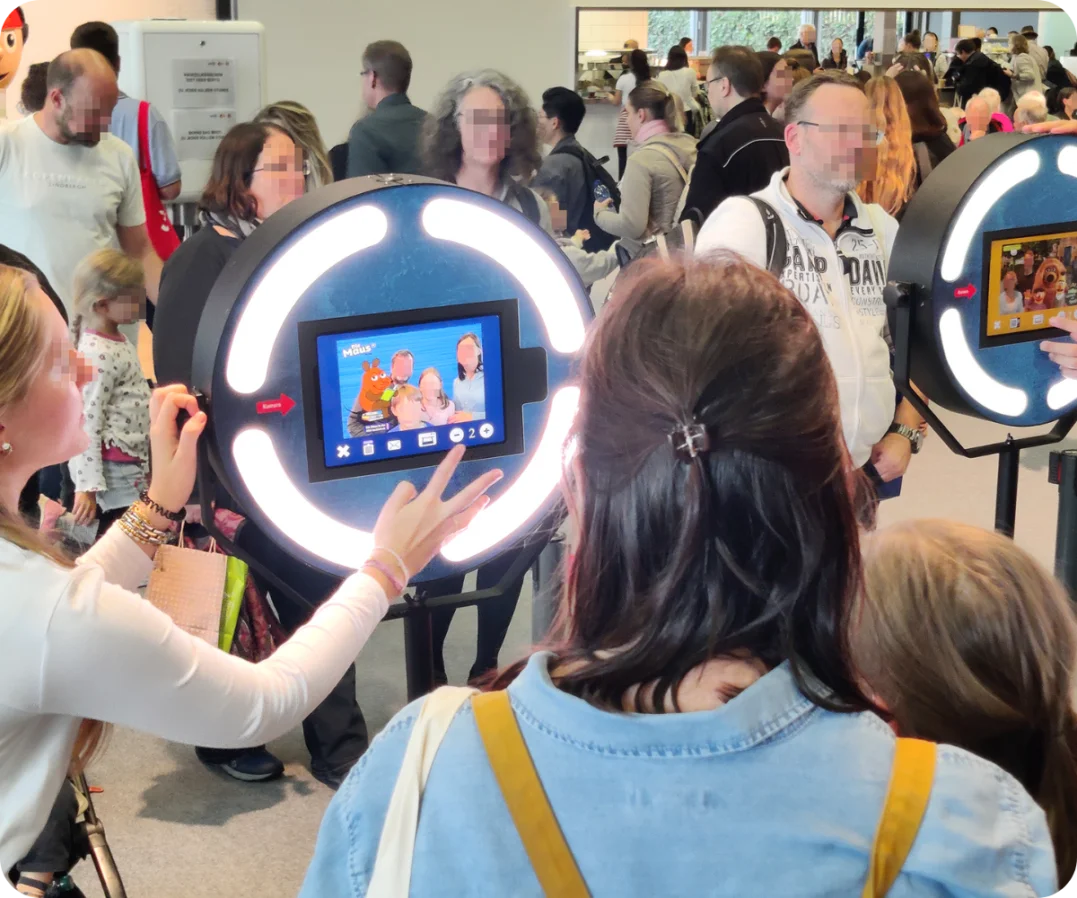 Besucherandrang an der Maus-Fotobox auf der Buchmesse Frankfurt Besucher stehen in einer Schlange, um Fotos an zwei Fotoboxen zu machen, die mit "Die Maus" Branding versehen sind. Auf den Bildschirmen sind beispielhafte Fotos mit den Charakteren zu sehen.