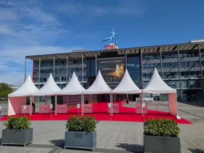 Außenansicht der MMC Studios in Köln bei strahlendem Sonnenschein. Ein roter Teppich führt unter weißen Zelten zum Eingang. Über dem Portal hängt ein großes Banner mit der Aufschrift 'Herzlich Willkommen - Der Deutsche Fernsehpreis 2025'. Auf dem Dach steht eine blaue Einhorn-Statue.