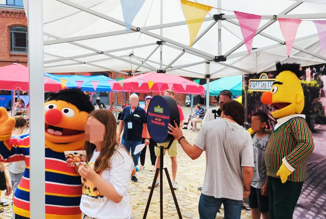Fotobox am HVV-Familienfest mit den Figuren der Sesamstraße Besucher, darunter Kinder und Jugendliche, posieren lachend an einer Fotobox auf einem Familienfest. Die Figuren Ernie und Bert aus der Sesamstraße sind ebenfalls anwesend. Im Hintergrund sind weitere Besucher und Stände unter Zelten zu sehen.