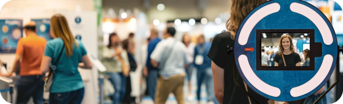 Detailaufnahme der betriebsbereiten Fotobox mit leuchtendem LED-Ring vor einem unscharfen, belebten Messe-Hintergrund. Suggeriert Einsatzbereitschaft für den nächsten Event.