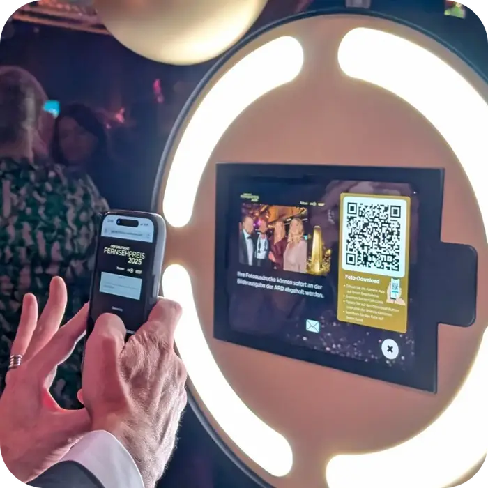 Nahaufnahme von Händen, die ein Smartphone halten. Der Gast scannt gerade einen QR-Code vom Display der Fotobox ab. Auf dem Handydisplay ist bereits die gebrandete Landingpage 'Der Deutsche Fernsehpreis 2025' mit Login-Maske zu sehen.