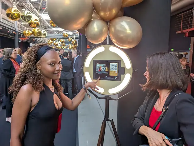 Zwei elegant gekleidete Gäste bedienen lachend den Touchscreen der AIR CORE Fotobox auf der Aftershow-Party. Im Hintergrund ist eine festliche Dekoration aus goldenen Luftballons zu sehen.